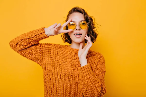 Close-up portrait of charming young woman with gently smile posing with peace sign in trendy glasses. Stylish girl in bright sweater having fun during photoshoot in studio..