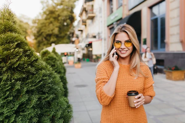 Laughing pretty lady talking on phone while walking by bushes with cup of tea. Outdoor portrait of ecstatic blonde woman in orange sweater spending time at the street and waiting friend with coffee..