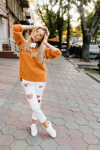 Full-length portrait of beautiful woman in white shoes and ripped pants playing with blonde hair. Photo of charming girl posing with legs crossed while spending time outdoor..