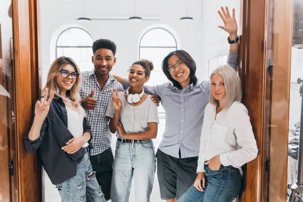 Slim african girl in jeans and white headphones standing in office with colleagues. European freelance female programmer having fun with friends and posing with pleasure..