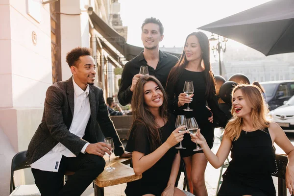 Joyful brunette man in black shirt looks away, holding glass of wine at party. Outdoor portrait of african guy in trendy jacket celebrating birthday with charming girls in cafe..