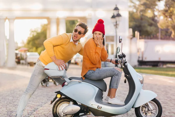 Elegant girl covers mouth with hands and looking away while sitting on scooter. Handsome guy in trendy dark sunglasses posing emotionally while his girlfriend learns to drive..