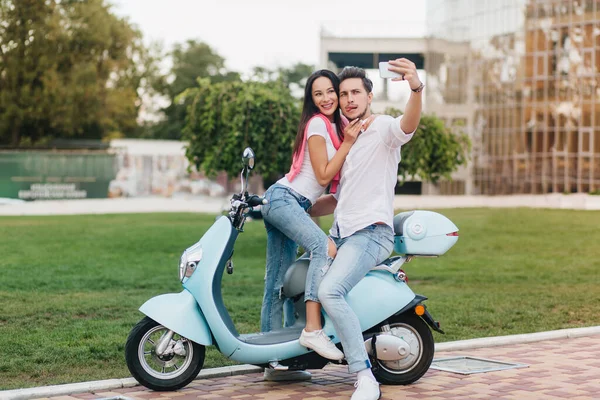 Caucasian man making funny faces while posing for selfie with brunette girl. Dark-haired guy sitting on scooter with young lady in white shoes and holding smartphone..