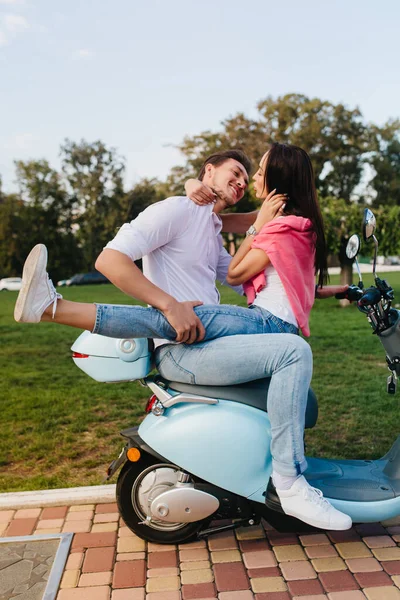Stylish smiling guy holding girlfriends legs while kissing her. Outdoor portrait of cute loving couple sitting on scooter together under blue sky..