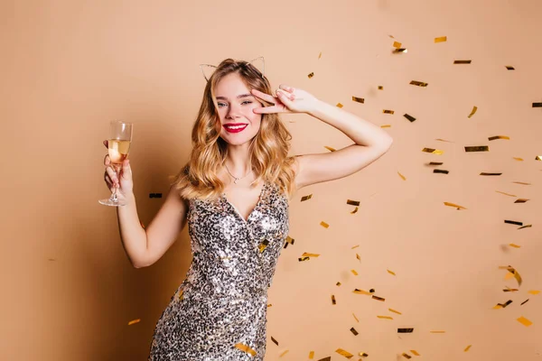 Stunning curly lovely girl holding glass of champagne and dancing with charming smile. Indoor portrait of elegant young lady in sparkle dress enjoying photoshoot under confetti..