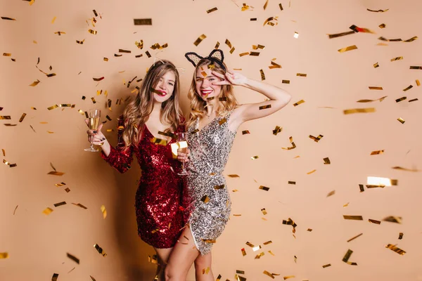 Funny curly girl in hair accessory expressing positive emotions under golden confetti. Indoor portrait of amazing female model in red sparkle clothes relaxing with best friend at hen-party..