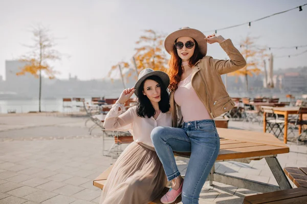 Blissful red-haired girl in jeans relaxing in street cafe with friend. Excited ladies posing together on autumn nature background..