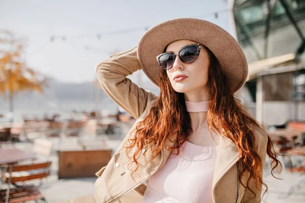 Pale woman with wavy dark hair looking up and touching her hat. Outdoor photo of chilling white girl in sunglasses posing on blur background in warm day..