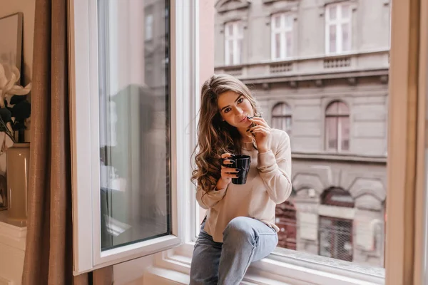 Romantic dark-haired girl spending time at home and drinking latte on sill. Portrait of dreamy caucasian young woman wears shirt and jeans posing next to window..