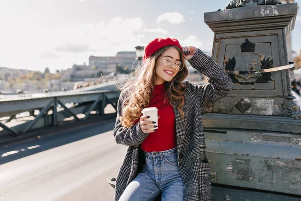 Fascinating shy woman in glasses drinking coffee on bridge in windy day. Outdoor portrait of ecstatic caucasian lady expressing happy emotions, holding cup of latte..