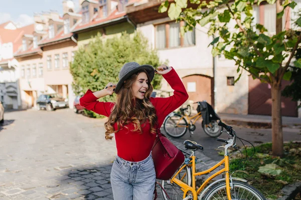 Sensual long-haired girl in red sweater having fun outdoor with bicycle. Portrait of stylish woman in vintage outfit dancing near green tree on city background..