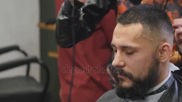 Nuevo peinado. Vista lateral del joven barbudo que se arregla en la peluquería con secador de pelo mientras está sentado en la silla en la barbería — Vídeos de Stock