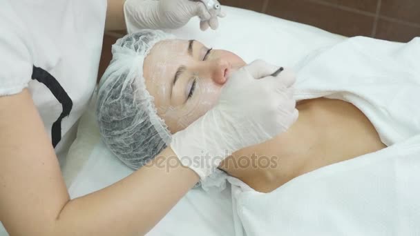 Κοντινό πλάνο της χέρι του cosmetologist εφαρμογή κρέμας μάσκα στο γυναικείο πρόσωπο. Κρατά βούρτσα. Η όμορφη νεαρή γυναίκα ξαπλωμένη με χαλάρωση — Αρχείο Βίντεο