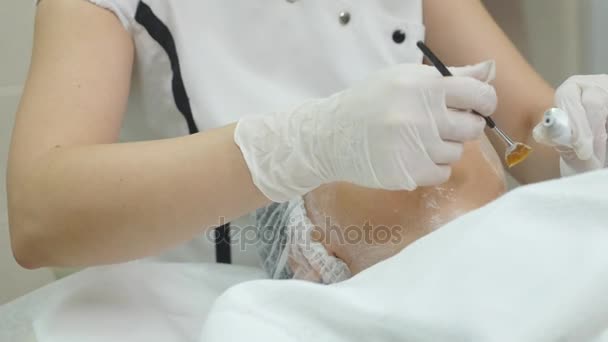 El primer plano de la mano del cosmetólogo que aplica la máscara de crema en la cara femenina. Está sosteniendo el cepillo. La hermosa joven está acostada con la relajación — Vídeos de Stock