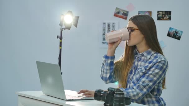 Красивая молодая женщина фотограф работает с ноутбуком и пить кофе в офисе — стоковое видео