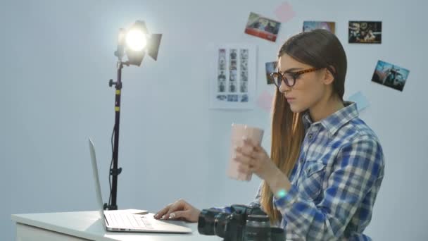 Linda joven fotógrafa trabajando con el ordenador portátil y tomando café en la oficina — Vídeos de Stock