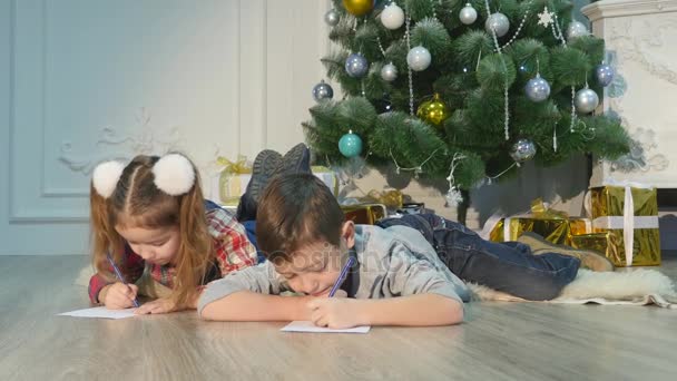Дети пишут письма Санта-Клаусу. Ребятам это весело. Мальчик и девочка лежат на полу возле рождественской елки. Дом празднично украшен . — стоковое видео