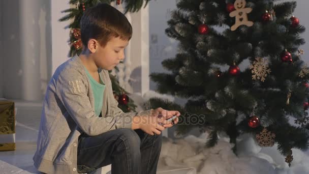 Niño jugando en el teléfono móvil, los niños y el teléfono inteligente cerca del árbol de Navidad — Vídeo de stock