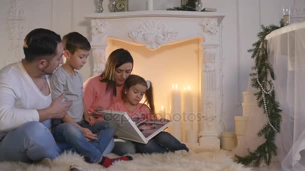 Familia citește cartea așezată pe canapea în fața șemineului în interiorul casei decorate de Crăciun — Videoclip de stoc