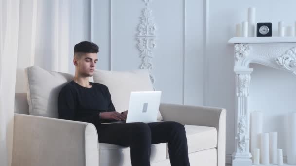 Hombre joven guapo sentado en el sofá y el uso de la computadora portátil en la habitación — Vídeos de Stock