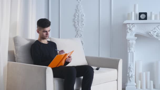 Joven guapo en casa escribiendo en el cuaderno, sentado en el sofá — Vídeos de Stock