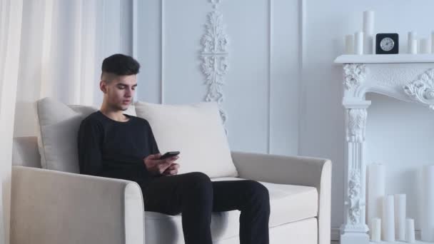 Tartózkodó touch otthon. Vidám fiatalember gazdaság mozgatható telefon, és mosolyogva, miközben feküdt a kanapén — Stock videók