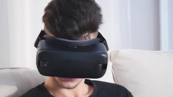 年轻的白人男子坐在沙发和有乐趣使用白色 Vr 耳机眼镜 — 图库视频影像