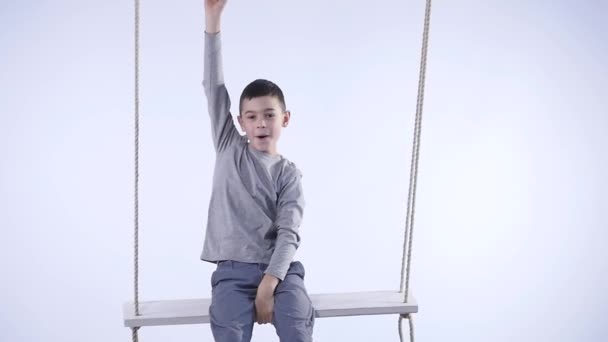 Радісний маленький хлопчик гойдається на гойдалці і жестикулює щастя ізольовано на білому тлі — стокове відео