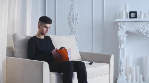 Bello giovanotto a casa a scrivere sul taccuino, seduto sul divano — Video Stock