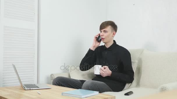 Junger Mann trinkt Kaffee und benutzt Laptop beim Telefonieren — Stockvideo