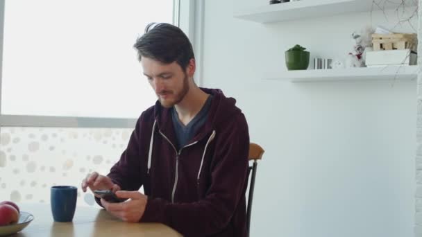 Jonge man met behulp van smartphone en het drinken van koffie door de tabel in de keuken — Stockvideo