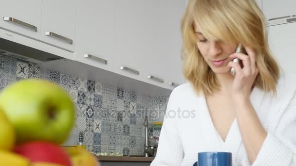 Sorrindo jovem mulher falando no telefone celular enquanto toma café da manhã em uma cozinha — Vídeo de Stock