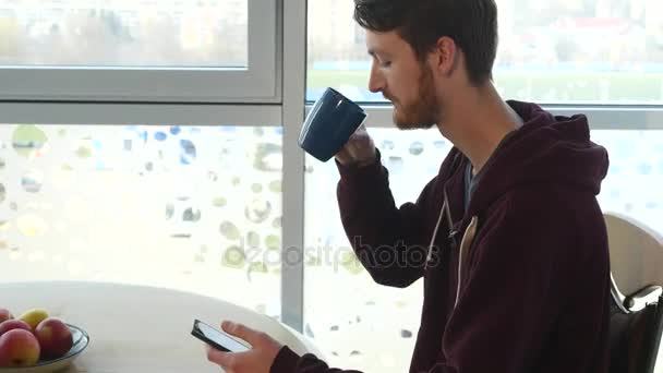 Ung man med smartphone och dricka kaffe av tabellen i kök — Stockvideo