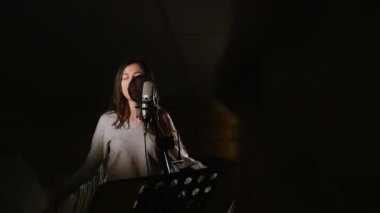 Güzel kız şarkı Studio mikrofona. Kız beyaz bir arka plan üzerinde kulaklık giyiyor. Stüdyo düşük ışık koşullarında.