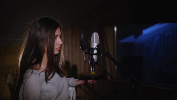 Menina bonita cantando no microfone do estúdio. A menina está usando fones de ouvido em um fundo branco. Na luz baixa do estúdio . — Vídeo de Stock