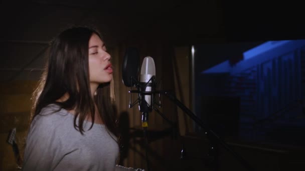 Pretty Girl Singing Into Studio Microphone. The girl is wearing headphones on a white background. In the Low light of the studio. — Stock Video