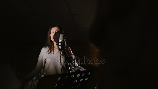 Pretty Girl Singing Into Studio Microphone. The girl is wearing headphones on a white background. In the Low light of the studio. — Stock Video