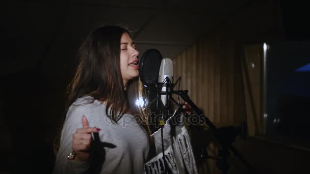 Pretty Girl Singing Into Studio Microphone. The girl is wearing headphones on a white background. In the Low light of the studio. — Stock Video