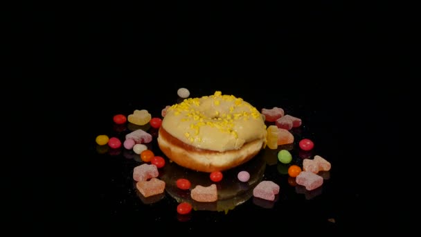 Gelbe lustige überraschte glasierte Donut mit Streusel, gestreifte Karamellbonbons, bunte Dragee mit Rosinen oder Erdnüssen im Inneren drehen sich auf schwarzem Hintergrund — Stockvideo