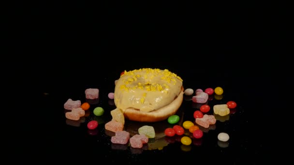 Amarillo divertido donut glaseado sorprendido con aspersiones, caramelos a rayas, dragee colorido con pasas o cacahuetes en el interior girar sobre fondo negro — Vídeo de stock