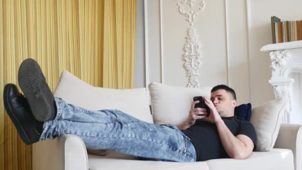 Hombre usando un teléfono inteligente, acostado en el sofá — Vídeos de Stock