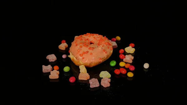 Πορτοκαλί αστείο έκπληκτος τζάμια ντόνατ με ψεκάζει, ριγέ καραμέλα καραμέλες, πολύχρωμο dragee με σταφίδες και φιστίκια στο εσωτερικό περιστρέφονται σε μαύρο φόντο — Φωτογραφία Αρχείου