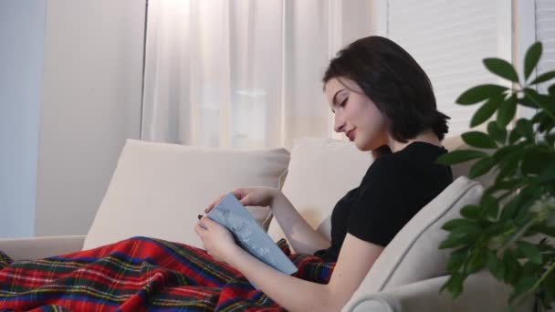 年轻漂亮的女人躺在沙发上，看书 — 图库视频影像