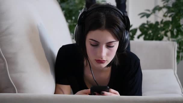 Красива приваблива молода жінка зі смартфоном лежить на дивані і слухає музику — стокове відео