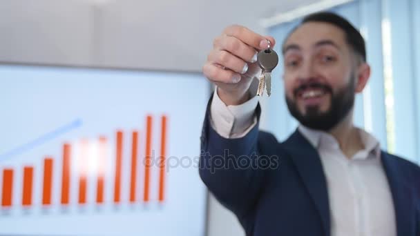 Schöner bärtiger Geschäftsmann im Anzug hält einen Autoschlüssel in der Hand, blickt in die Kamera und lächelt dabei — Stockvideo
