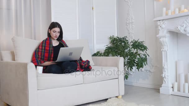 Junge Frau mit Laptop trinkt Kaffee, während sie auf der Couch im Wohnzimmer sitzt — Stockvideo