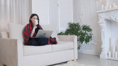 Oturma odasında kanepede otururken kahve içme dizüstü kullanan genç kadın