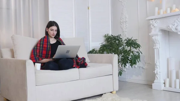 Junge Frau mit Laptop trinkt Kaffee, während sie auf der Couch im Wohnzimmer sitzt — Stockfoto