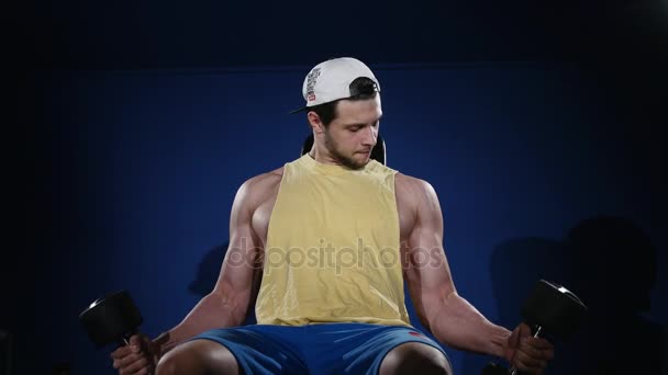 Joven atleta musculoso construido haciendo ejercicio en un gimnasio, sentado en una máquina de levantamiento de pesas y levantando dos pesas — Vídeo de stock