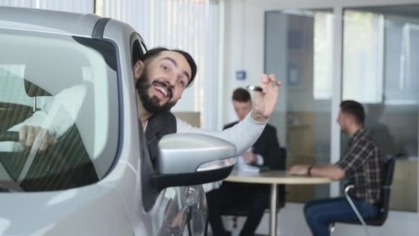 Besök i showroom. Ung man får håll bilnycklar, tittar på kameran och ler, sitter i sin nya bil — Stockvideo